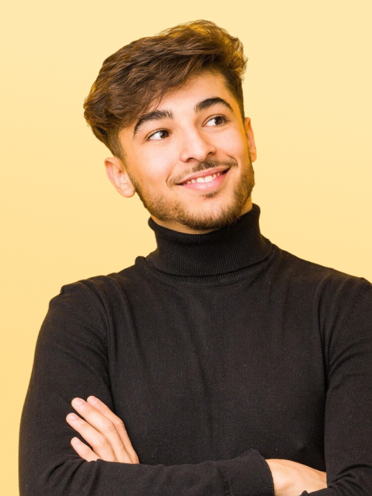 Tânăr cu păr șaten și barbă, purtând un pulover negru cu guler înalt, zâmbind și privind în lateral cu brațele încrucișate, pe un fundal galben.
