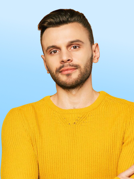 Bărbat cu barbă și păr scurt, purtând un pulover galben, pe un fundal albastru gradient.