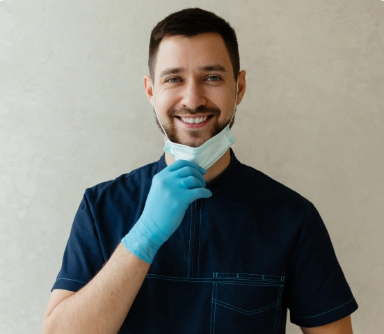 Bărbat zâmbitor cu păr scurt închis la culoare și barbă, îmbrăcat în scrubs medicale albastre și mănuși, dând jos masca chirurgicală.