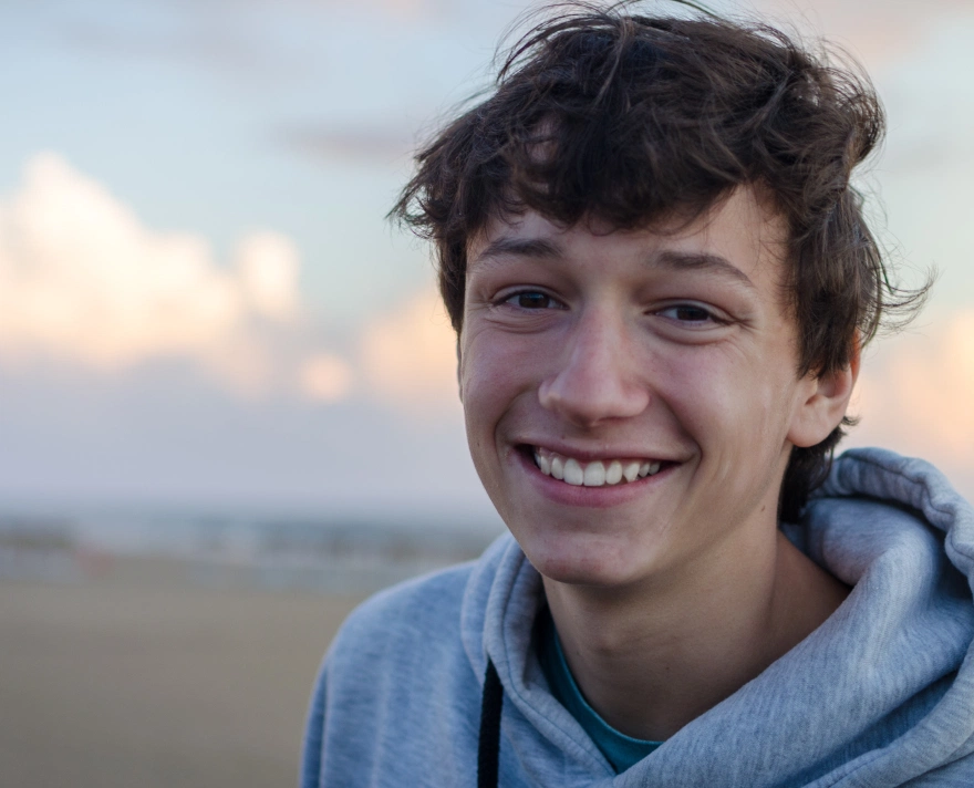 Adolescent zâmbitor cu păr ciufulit și hanorac gri, pe un fundal exterior estompat la apus de soare.