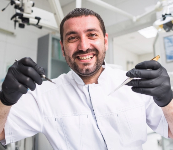 Dentist zâmbitor purtând mănuși negre și halat alb, ținând instrumente dentare într-o clinică.